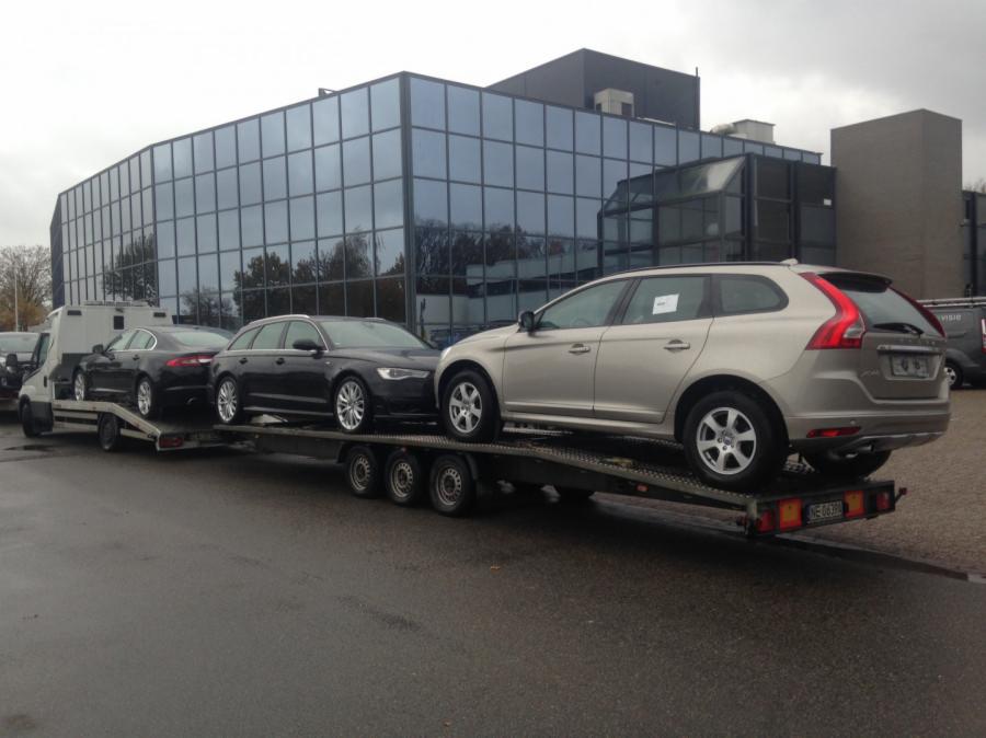 Laweta przewozi trzy samochody osobowe: srebrne Volvo XC60, czarne Audi A6 Avant i czarnego Jaguara XF na tle nowoczesnego biurowca z szklaną fasadą.