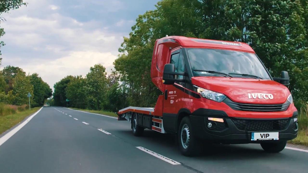 Czerwona laweta Iveco marki SPOJKAR z tablicą rejestracyjną VIP, jadąca asfaltową drogą wśród zieleni drzew.