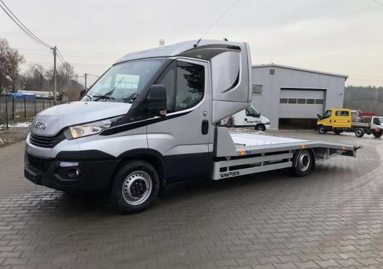 Srebrna laweta marki Iveco z platformą załadunkową na kostce brukowej w Kamieniu Pomorskim, widoczna z boku, z budynkiem w tle i innymi pojazdami.