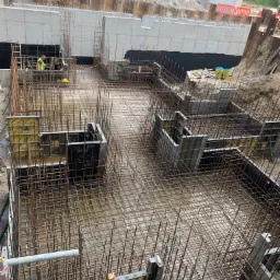 Budowa fundamentów: widok z góry na szkielet z prętów stalowych i szalunki, z pracownikiem w kamizelce odblaskowej w tle, teren budowy z ziemią i konstrukcją stalową w tle.