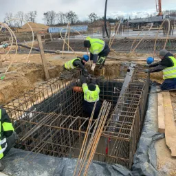 Pracownicy w odblaskowych kamizelkach montują szkielet z prętów zbrojeniowych w wykopie zabezpieczonym folią, widoczna drabina i pompa do betonu w tle.