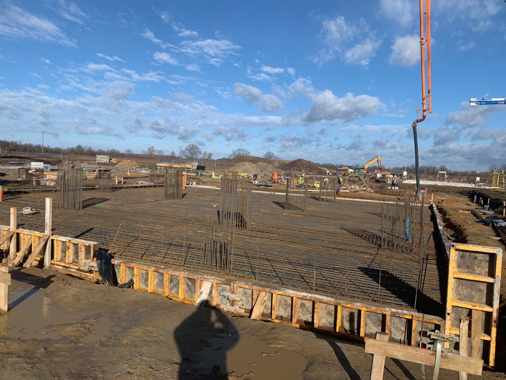 Przygotowanie zbrojenia i szalunków pod fundamenty na placu budowy, widoczna pompa do betonu i pracownicy w oddali.