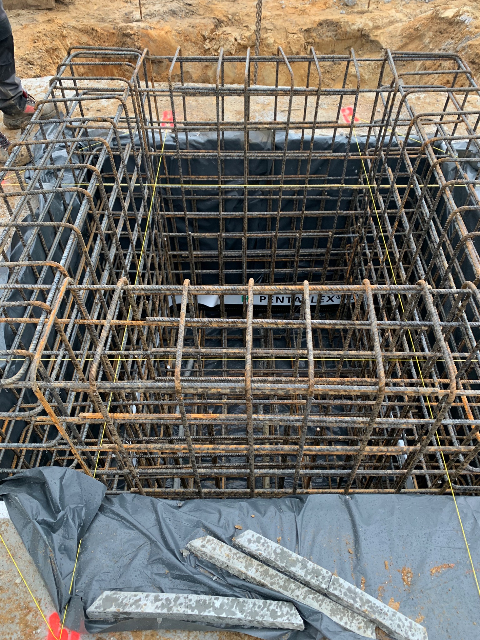 Klatka z prętów zbrojeniowych w wykopie zabezpieczonym czarną folią, widoczne żółte sznurki i fragment stopy pracownika w lewym górnym rogu.