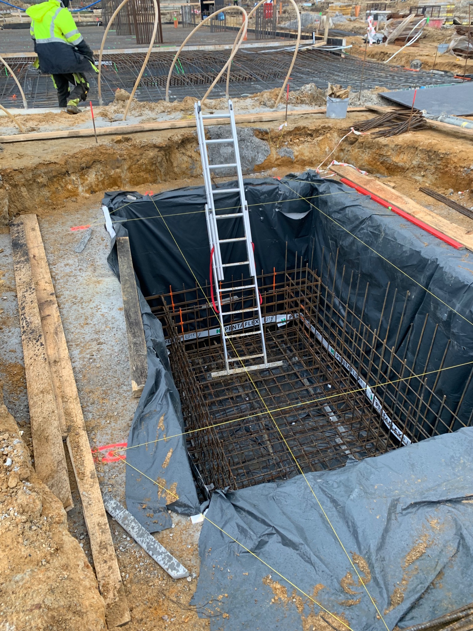 Wykonanie szkieletu zbrojeniowego w wykopie fundamentowym zabezpieczonym folią, widoczna drabina i pracownik w odblaskowej kurtce w tle.