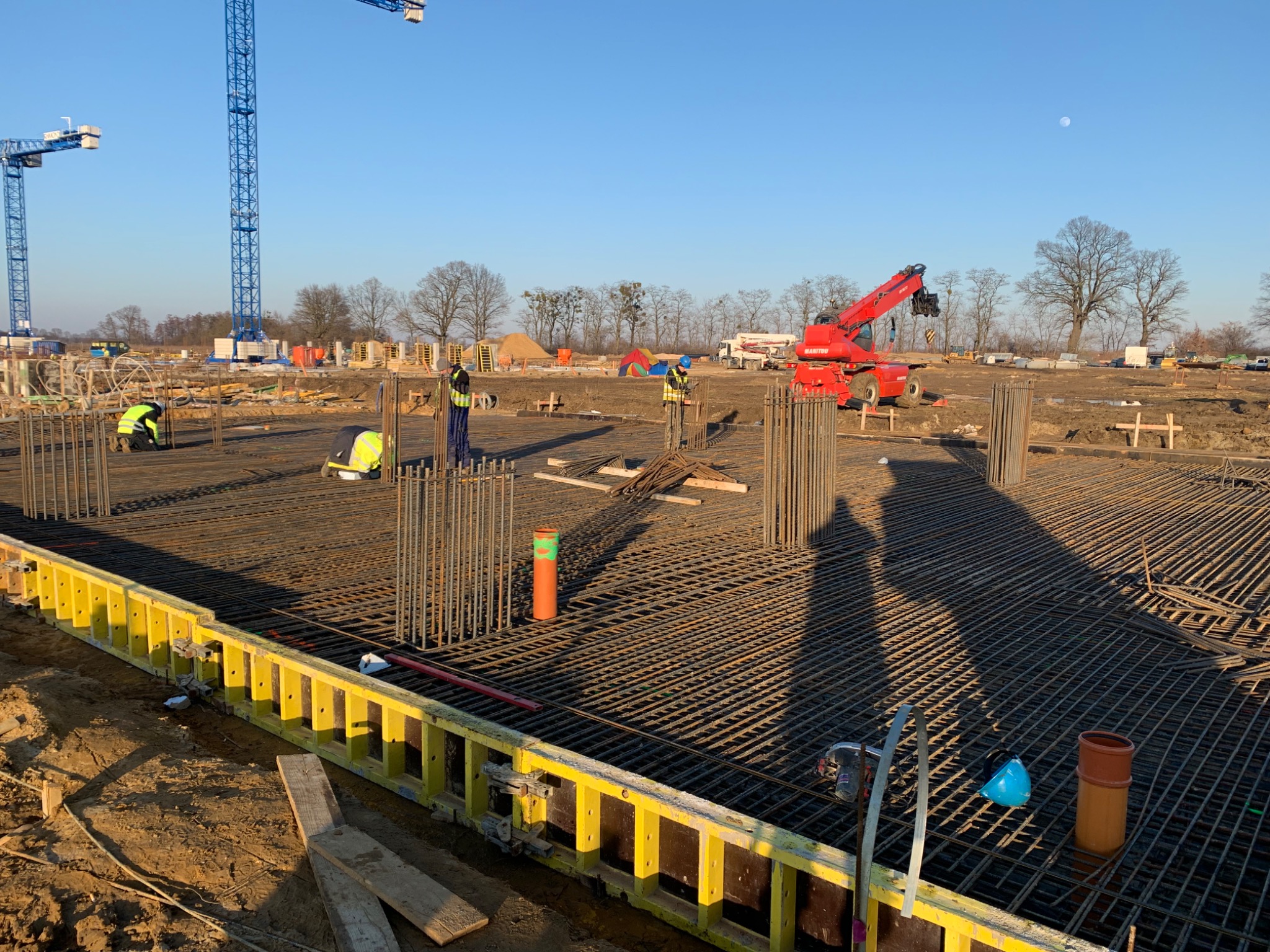 Przygotowanie zbrojenia fundamentów na placu budowy; widoczna siatka z prętów, pionowe słupy zbrojeniowe, żółte szalunki, pracownicy w odblaskowych kamizelkach, dźwigi w tle i czerwony podnośnik...