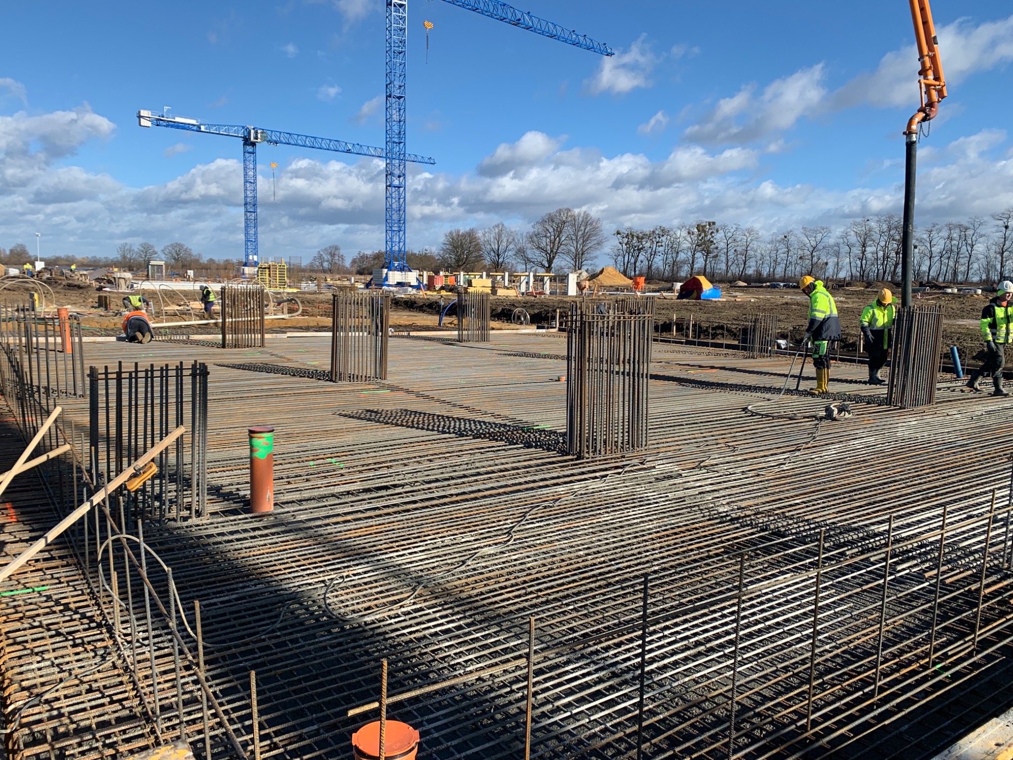Siatka zbrojeniowa przygotowana pod wylewkę fundamentów, widoczne pionowe pręty zbrojeniowe, pracownicy w żółtych kamizelkach i kaskach oraz dźwigi budowlane w tle.