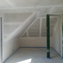 Pokój na poddaszu w trakcie wykańczania płytami kartonowo-gipsowymi, widoczne szpachlowane łączenia, zabezpieczone taśmą malarską krawędzie i pionowa drewniana podpora stropu.