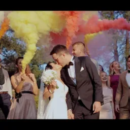 Para młoda całuje się na tle kolorowych dymów odpalonych przez gości weselnych, w tle drzewa i biały samochód.