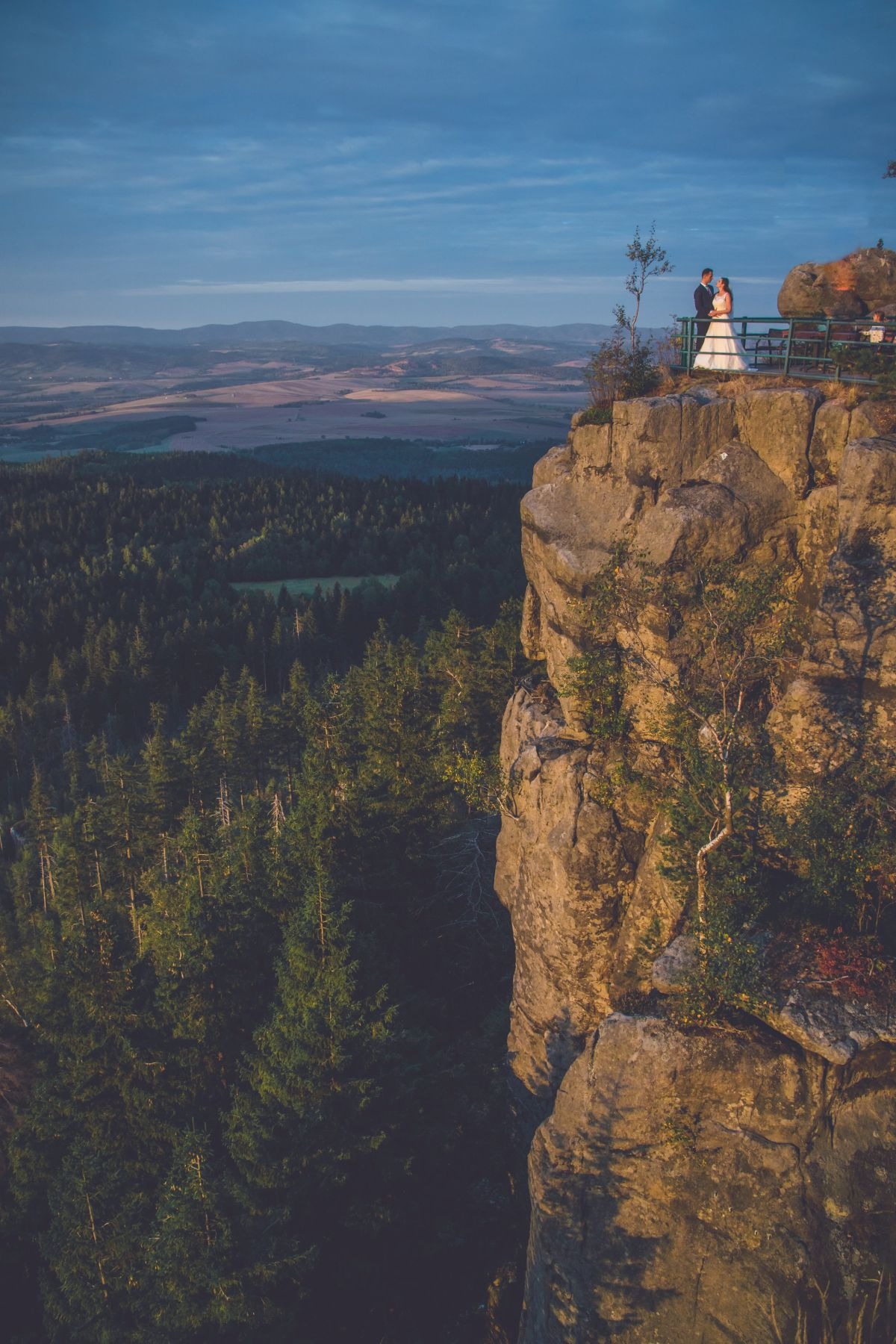 Para młoda stoi na platformie widokowej na szczycie skalnego urwiska, z rozległą panoramą lasów i pól w tle, uchwycona w ciepłym świetle zachodzącego słońca.