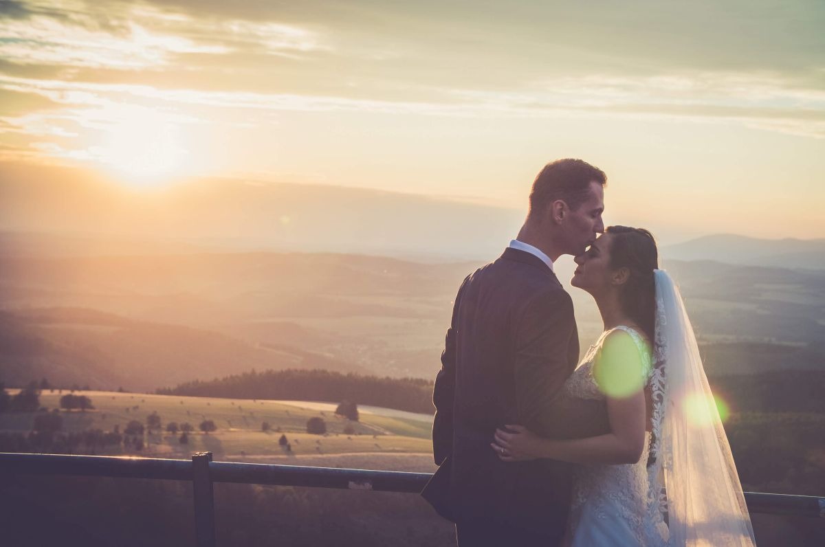 Para młoda na tle zachodzącego słońca i rozległego krajobrazu górskiego, panna młoda w koronkowej sukni, pan młody w ciemnym garniturze, on całuje ją w czoło.