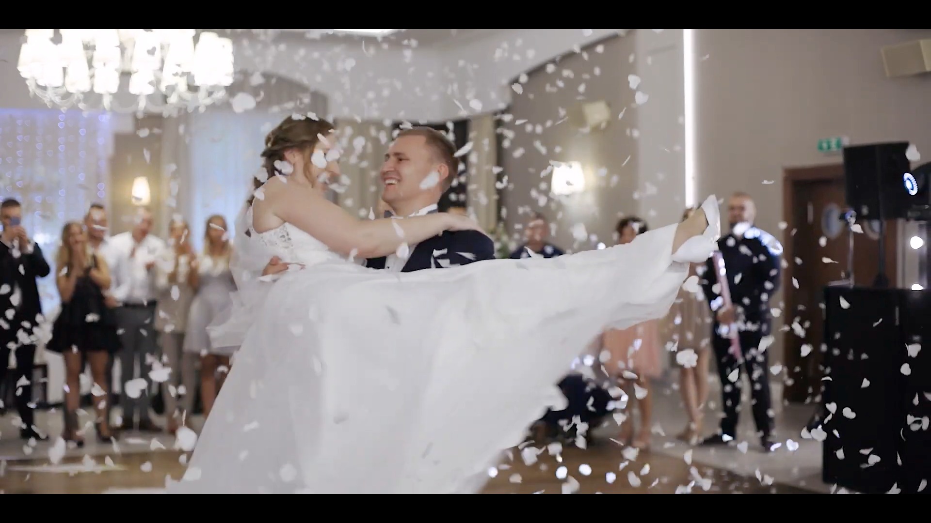 Uśmiechnięty pan młody trzyma na rękach pannę młodą podczas pierwszego tańca, w powietrzu opadają białe płatki konfetti, w tle goście i dekoracje sali.