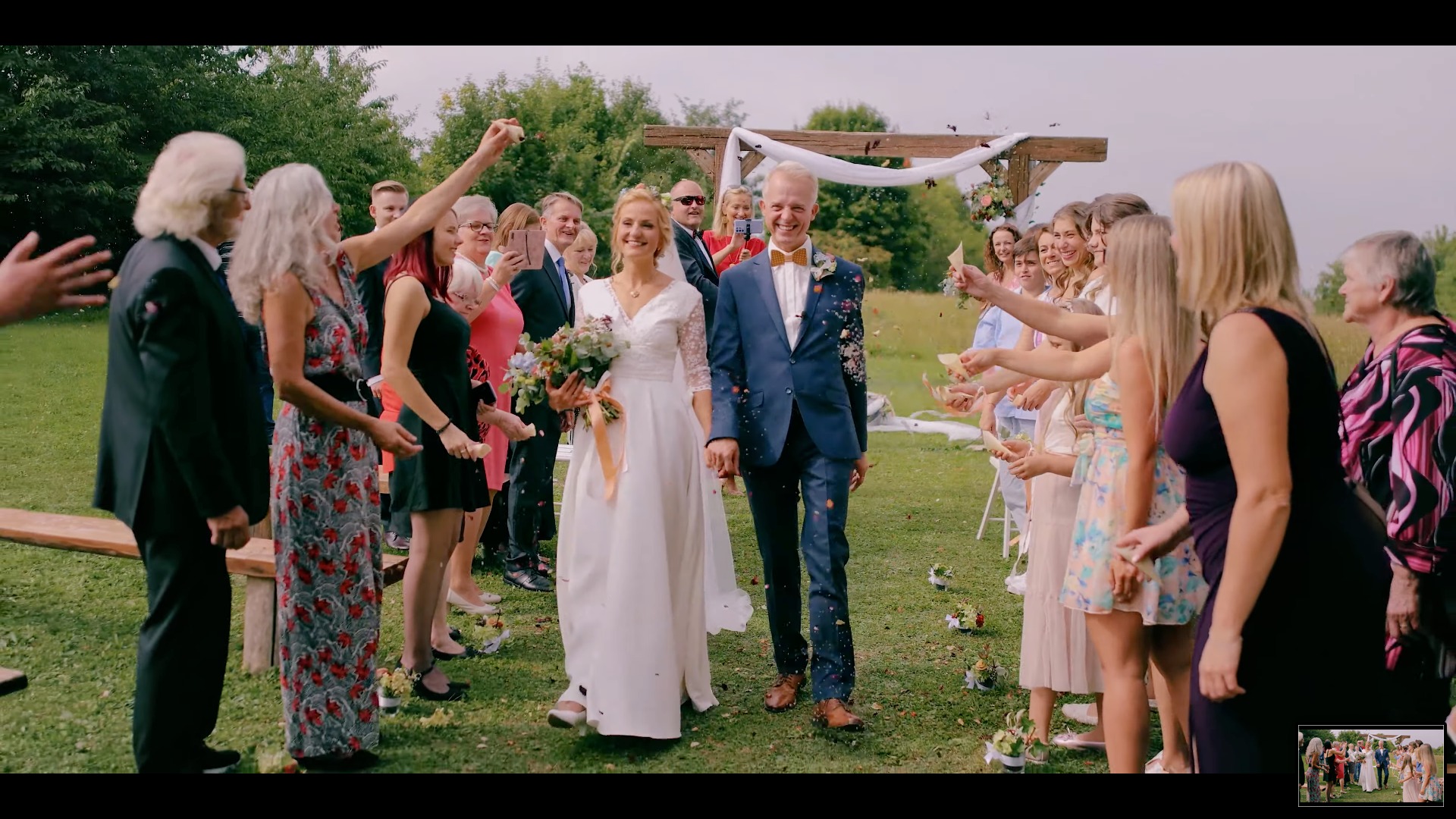 Para młoda, uśmiechnięta, przechodzi pod drewnianą bramą, obsypywana konfetti przez gości weselnych na zielonym trawniku.