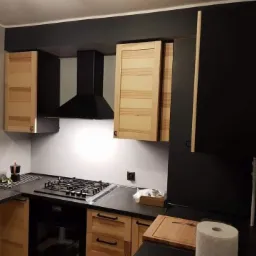 Kuchnia po remoncie: jasne drewniane szafki wiszące kontrastują z czarnymi elementami, takimi jak okap i pas nad szafkami, płyta gazowa wkomponowana w czarny blat.