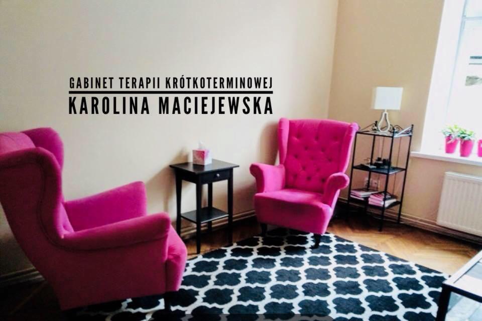 Jasny gabinet z dwoma różowymi fotelami typu uszak, czarno-białym dywanem w marokański wzór, stolikiem pomocniczym i napisem na ścianie 'Gabinet terapii krótkoterminowej Karolina Maciejewska'.