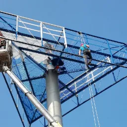 Dwóch pracowników na wysokościach przy konstrukcji reklamowej: jeden w koszu podnośnika, drugi zabezpieczony linami, obaj w kaskach, na tle błękitnego nieba.