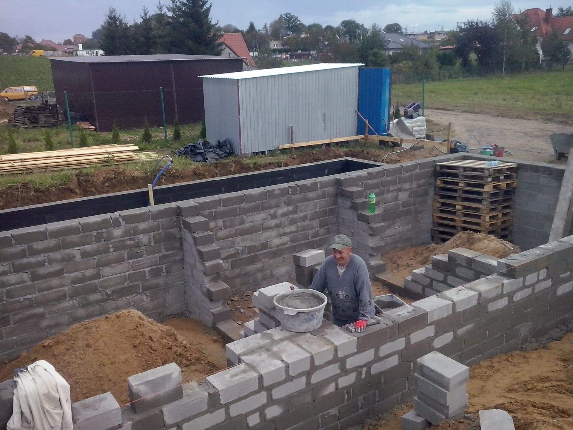 Budowa fundamentów z bloczków betonowych: murarz w trakcie pracy, widoczny fragment wznoszonej ściany fundamentowej, zaprawa w wiadrze, palety z bloczkami w tle, teren budowy z widocznymi...