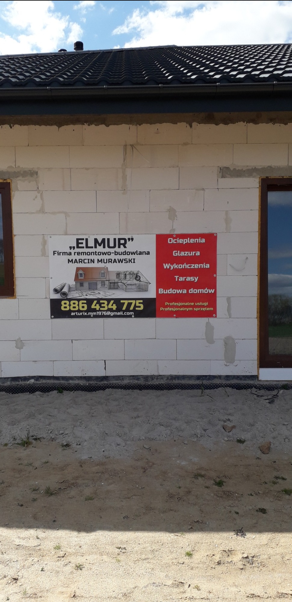 Ściana budynku w trakcie budowy z białych bloczków, na której umieszczone są dwie tablice reklamowe firmy budowlanej 'Elmur' oferującej usługi budowlane, ociepleniowe i wykończeniowe, widoczne okno...