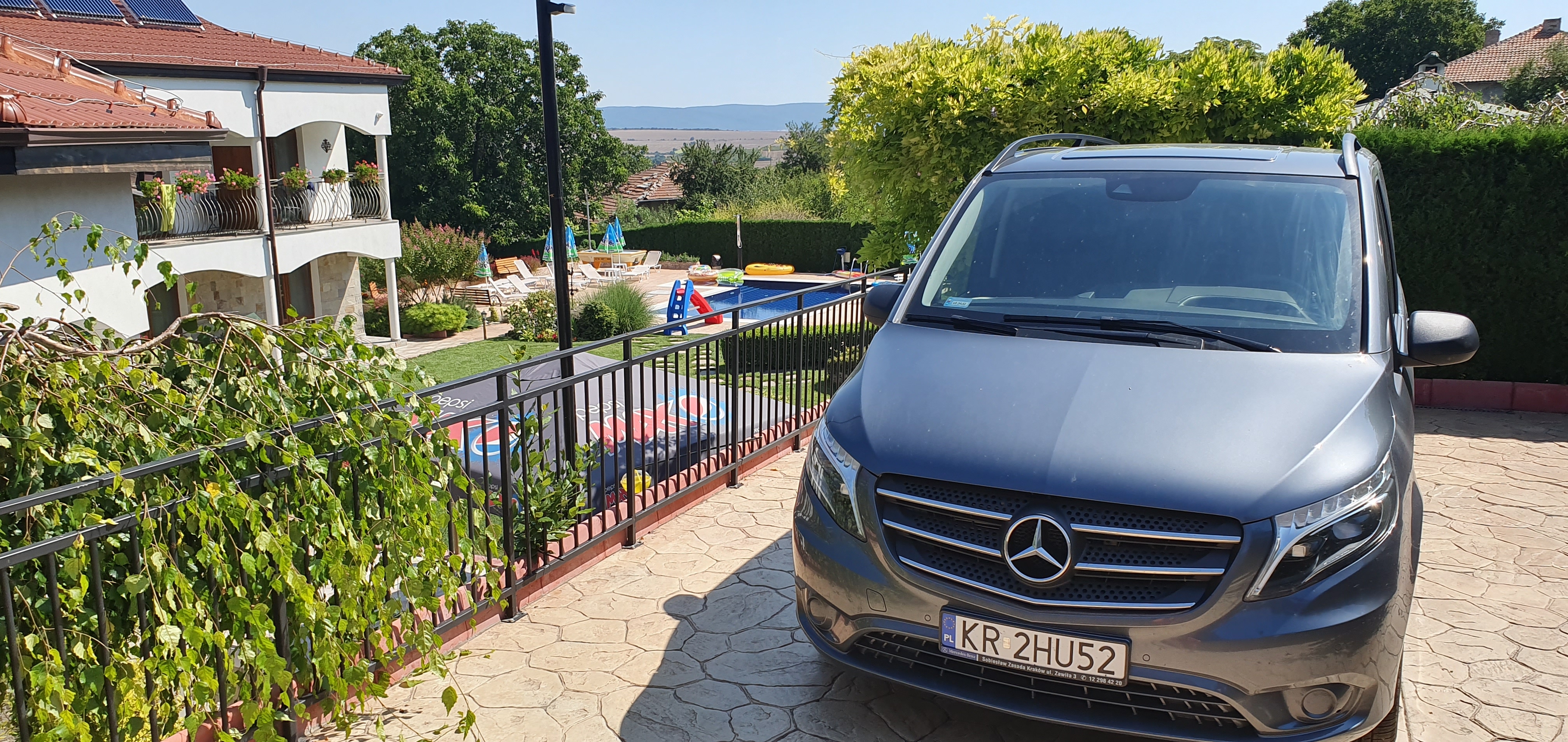 Szary van Mercedes-Benz z polską tablicą rejestracyjną na tle luksusowego ogrodu z basenem i białym domem z balkonami
