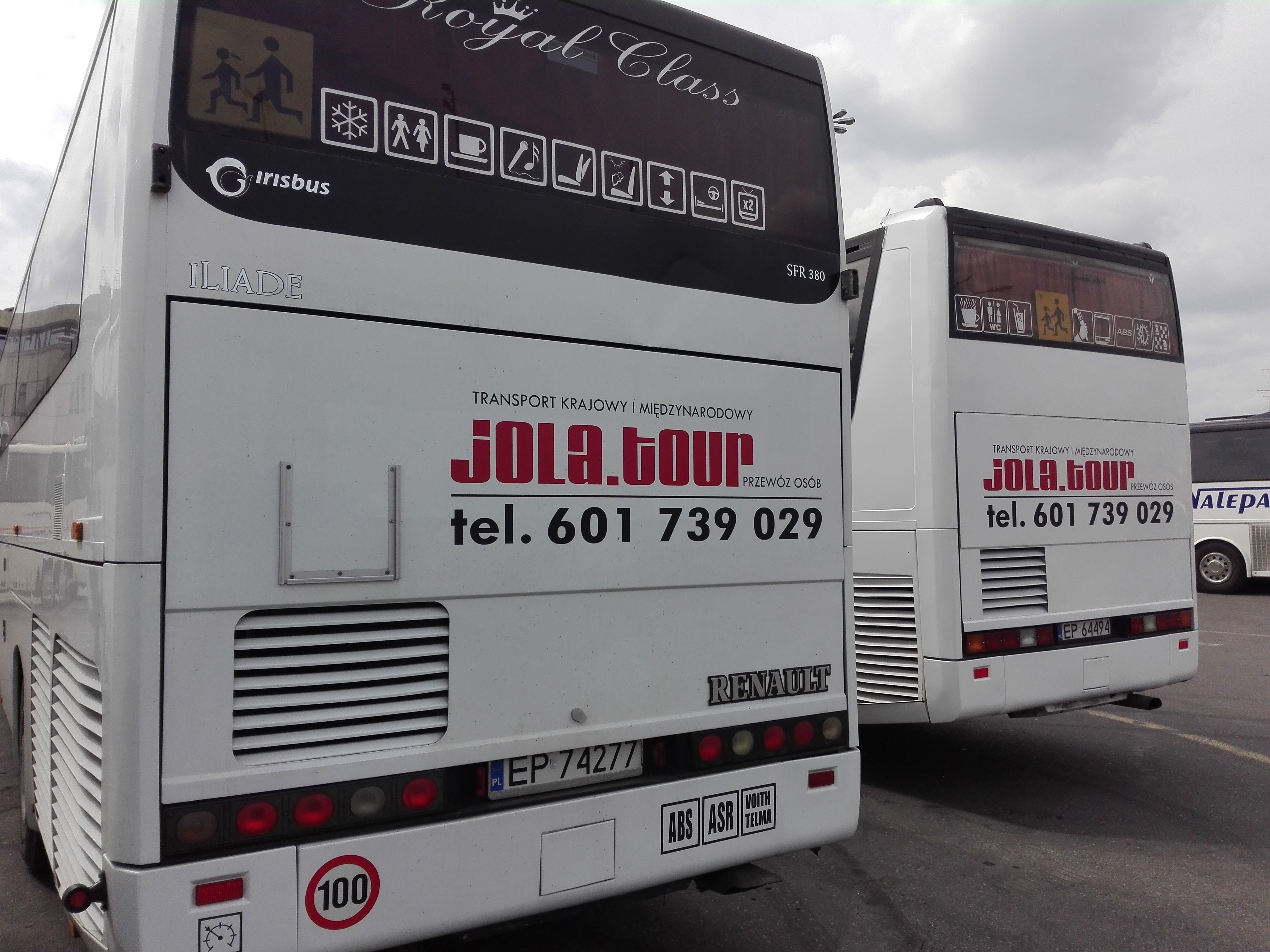 Dwa białe autokary firmy JOLA.tour z numerami telefonów i oznaczeniami wyposażenia, zaparkowane na zewnątrz.