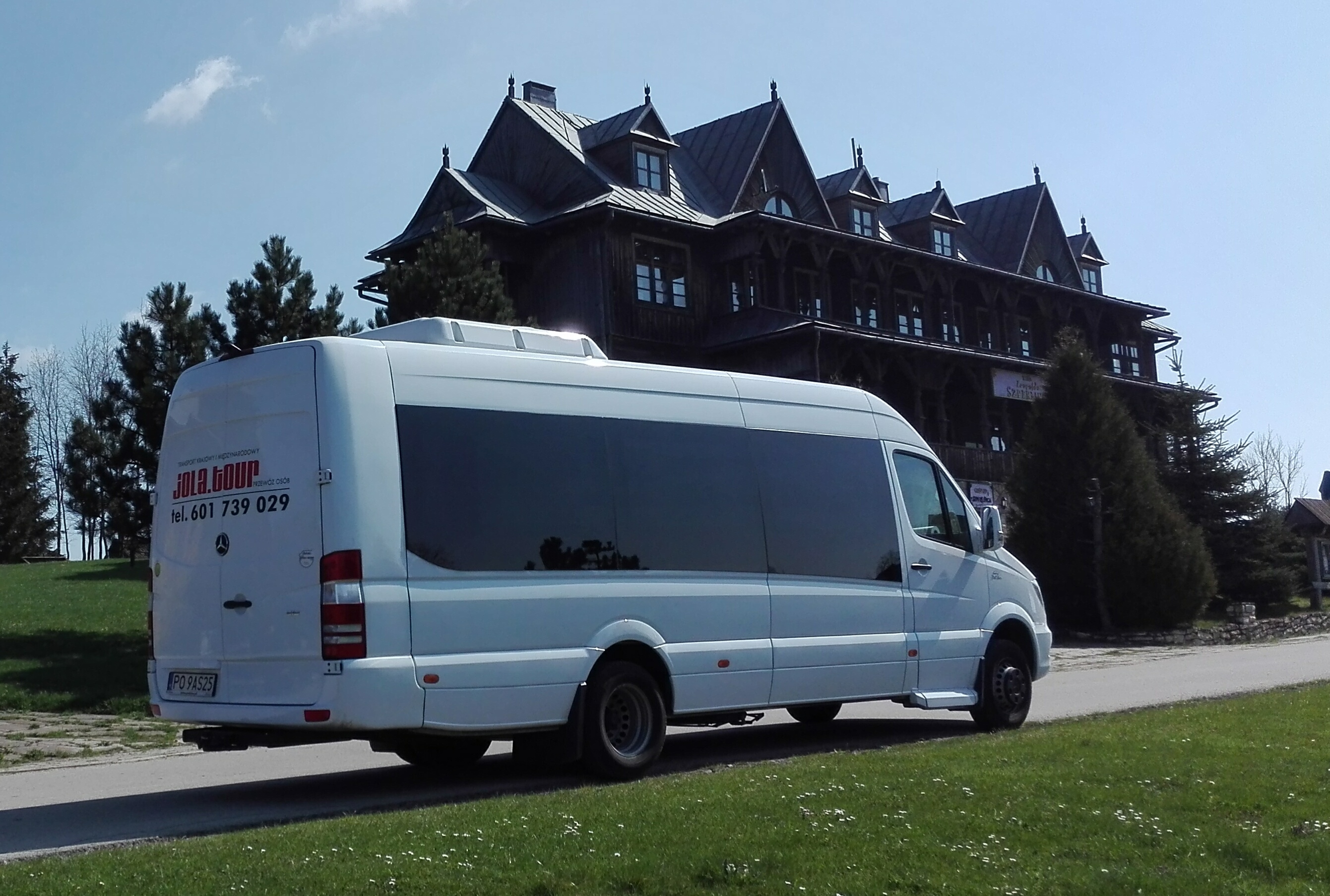 Biały bus Mercedes-Benz z napisem 'JOLA.tour' i numerem telefonu, zaparkowany na tle drewnianego budynku w stylu zakopiańskim.