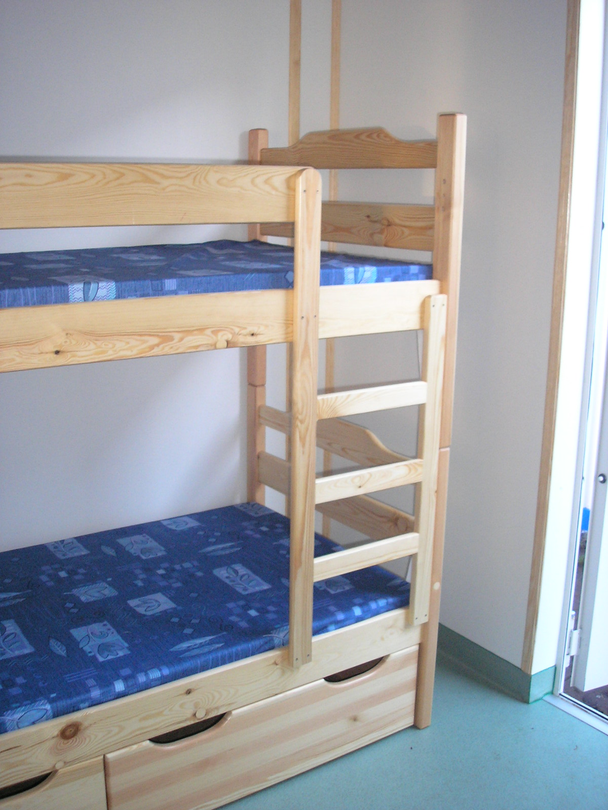 Drewniane łóżko piętrowe z niebieskimi materacami w pokoju z jasnymi ścianami i podłogą, widoczne szuflady pod dolnym łóżkiem i drabinka.