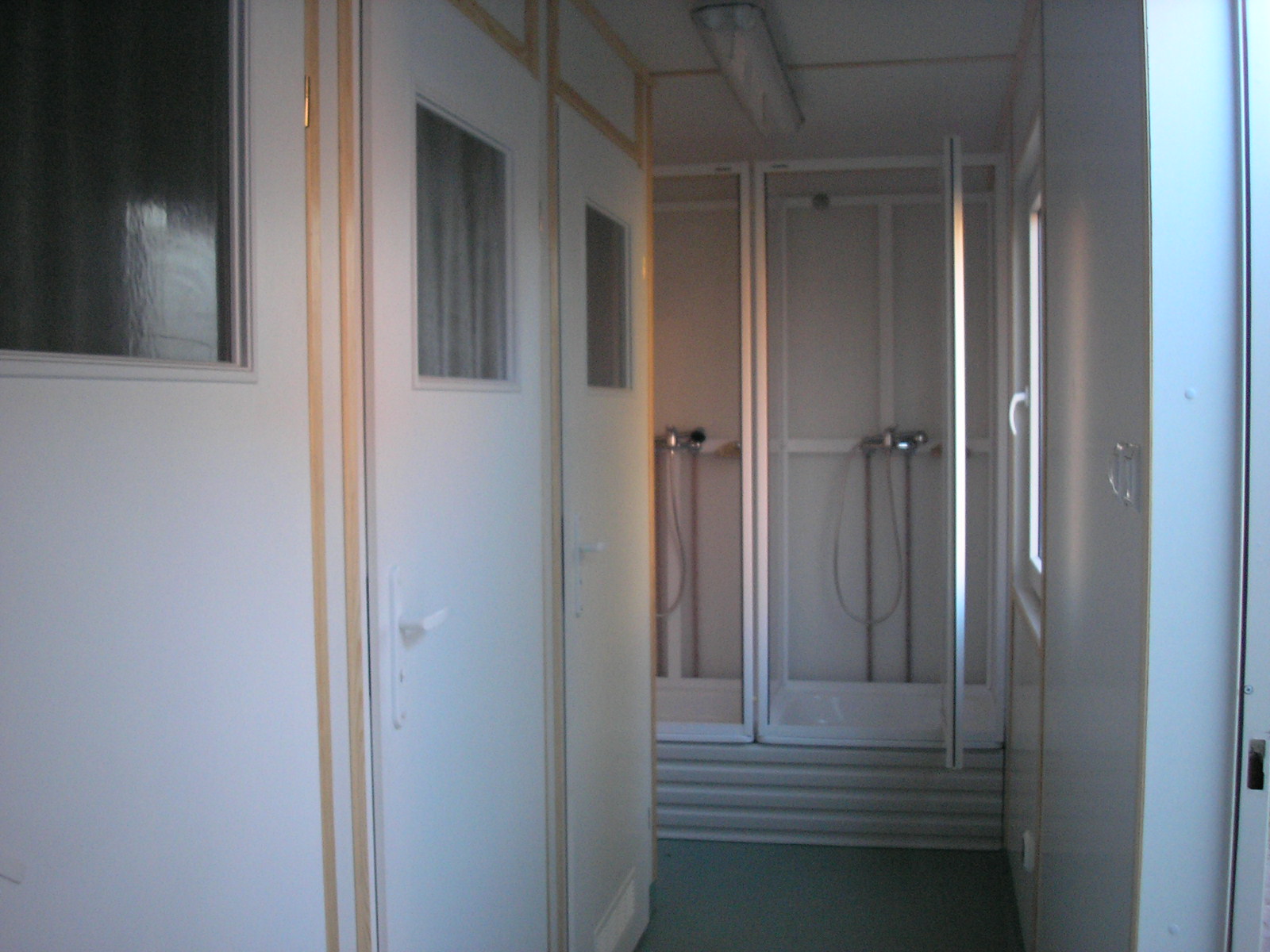 Wnętrze kontenera sanitarnego z kabinami prysznicowymi i białymi drzwiami z oknami, widok z korytarza, oświetlenie sufitowe.