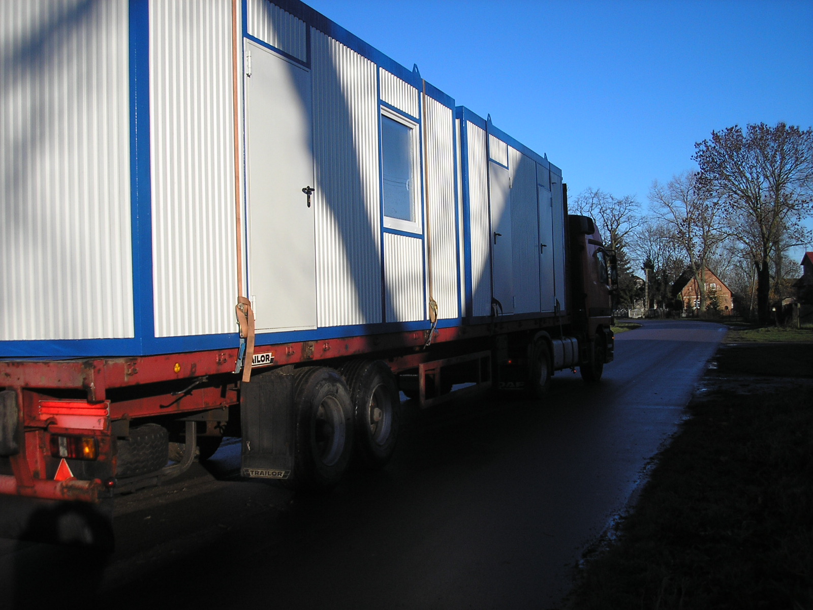 Transport kontenerów biurowych na naczepie ciężarówki po asfaltowej drodze w słoneczny dzień.