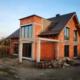 Surowy, murowany dom w trakcie budowy z widocznymi rusztowaniami, częściowo położonym dachem z dachówki, niezabudowanym tarasem i stertą drewna na placu budowy.