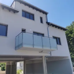 Szklana balustrada na balkonie nowoczesnego budynku z widokiem na błękitne niebo, z kolumnami podtrzymującymi balkon i widocznym fragmentem dachu.