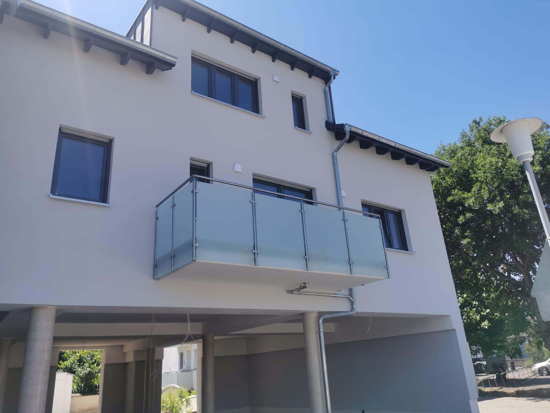 Szklana balustrada na balkonie nowoczesnego budynku z widokiem na błękitne niebo, z kolumnami podtrzymującymi balkon i widocznym fragmentem dachu.