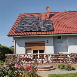 Panele słoneczne na dachu domu jednorodzinnego z czerwoną dachówką, widok z ogrodu w słoneczny dzień.