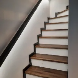 Nowoczesne schody z drewnianymi stopniami i czarną metalową balustradą, oświetlone punktowo od góry.