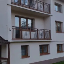 Dwupiętrowy budynek z balkonami zabezpieczonymi metalowymi balustradami z dekoracyjnymi elementami, widoczne okna z drewnianymi ramami, trawnik przed budynkiem.