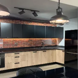 Nowoczesna kuchnia z ceglaną ścianą, czarnymi szafkami górnymi i drewnopodobnymi dolnymi, oświetlona lampami wiszącymi w stylu industrialnym.