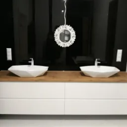 Łazienka z dwoma umywalkami o geometrycznym kształcie, na drewnianym blacie i białej szafce, na tle czarnych płytek i ozdobnego lustra.