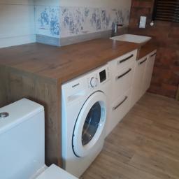 Kristol - Nowoczesna łazienka z zabudową na wymiar: drewniany blat nad pralką i szafkami, umywalka nablatowa, toaleta kompaktowa, płytki ścienne z motywem kwiatowym i grzejnik drabinkowy na ścianie z cegły.