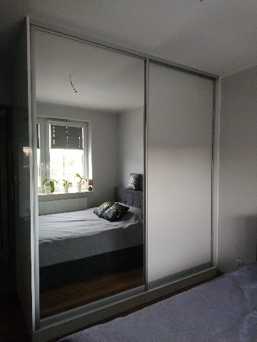 Szafa przesuwna z lustrem i matową szybą, widok na sypialnię z łóżkiem i oknem.