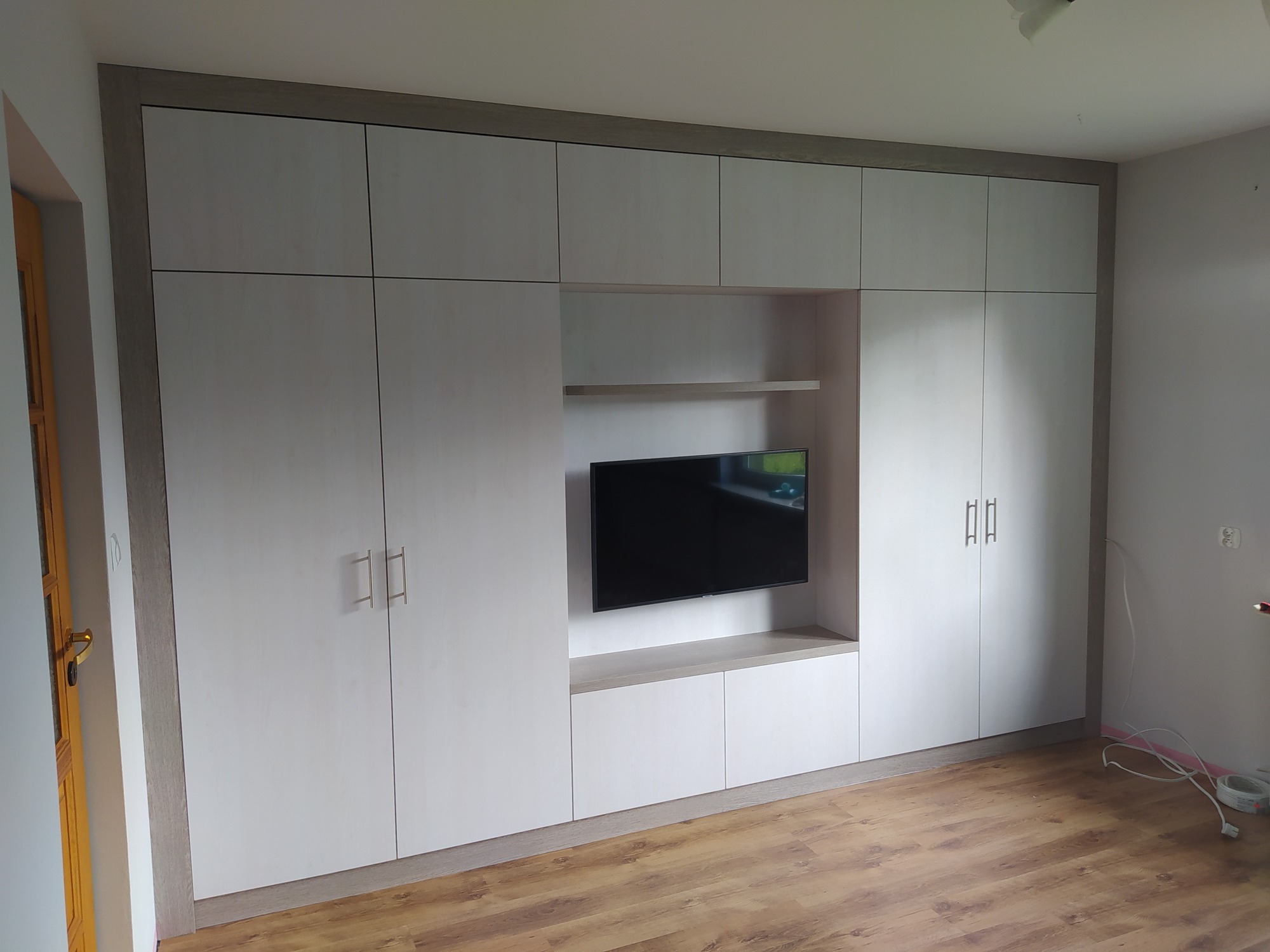 Biała zabudowa meblowa z szafkami i wnęką na telewizor, widoczne kable na podłodze