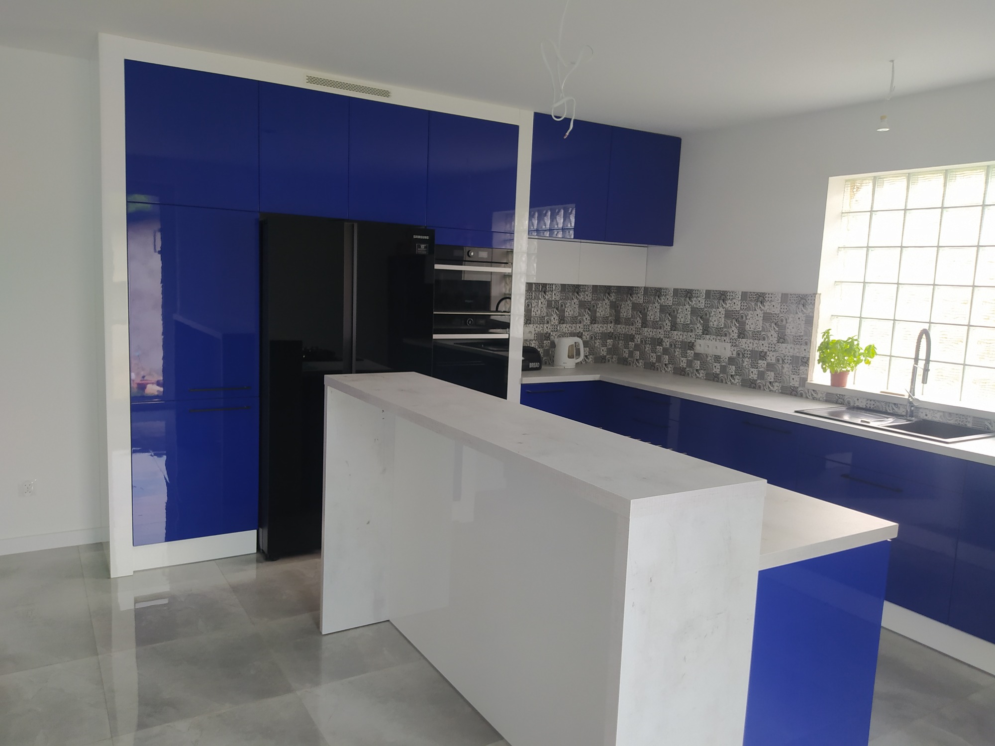 Nowoczesna kuchnia z błyszczącymi, kobaltowymi szafkami, białym blatem i wyspą kuchenną, czarną lodówką oraz mozaiką nad blatem przy oknie.
