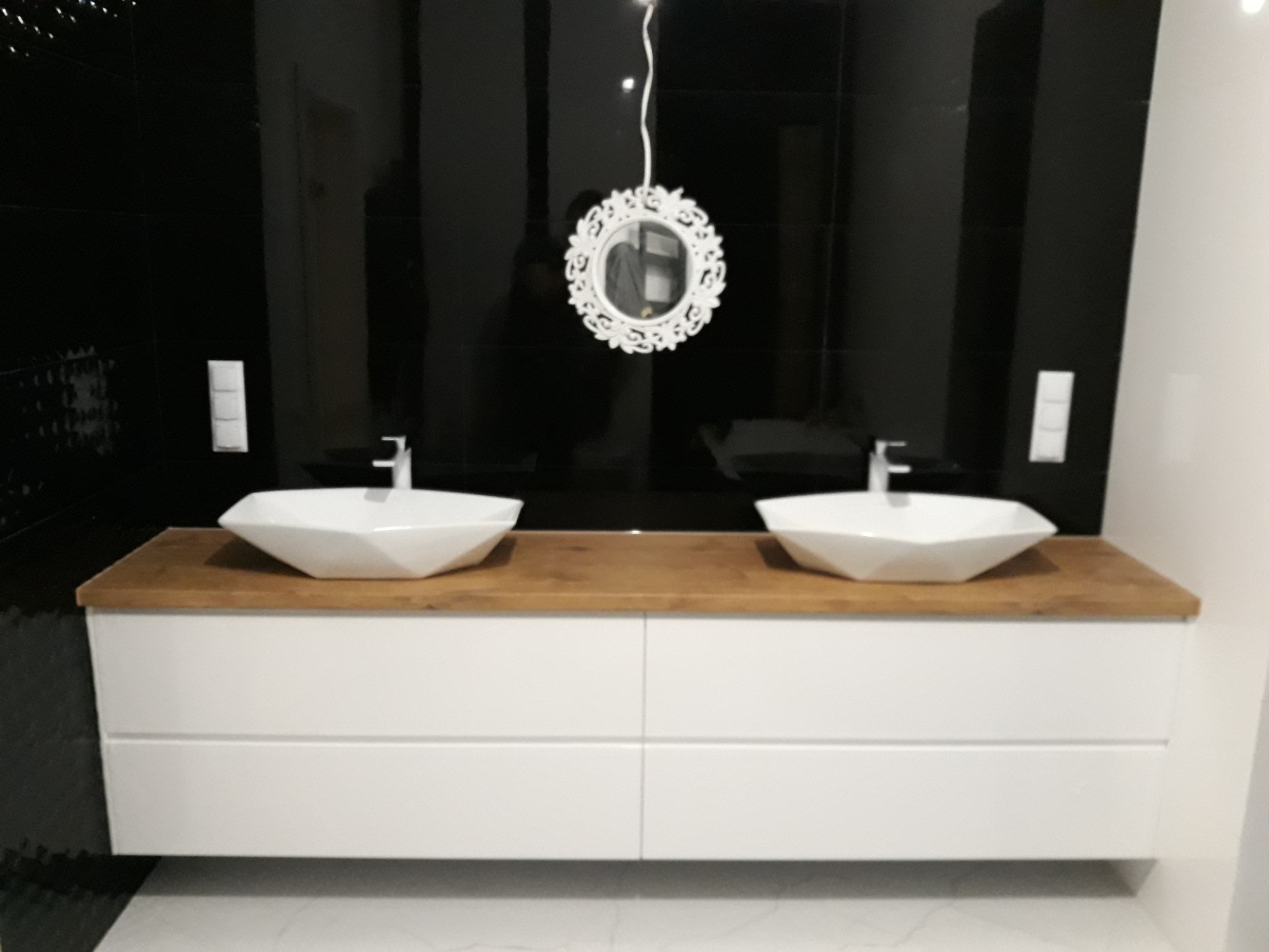 Łazienka z dwoma umywalkami o geometrycznym kształcie, na drewnianym blacie i białej szafce, na tle czarnych płytek i ozdobnego lustra.