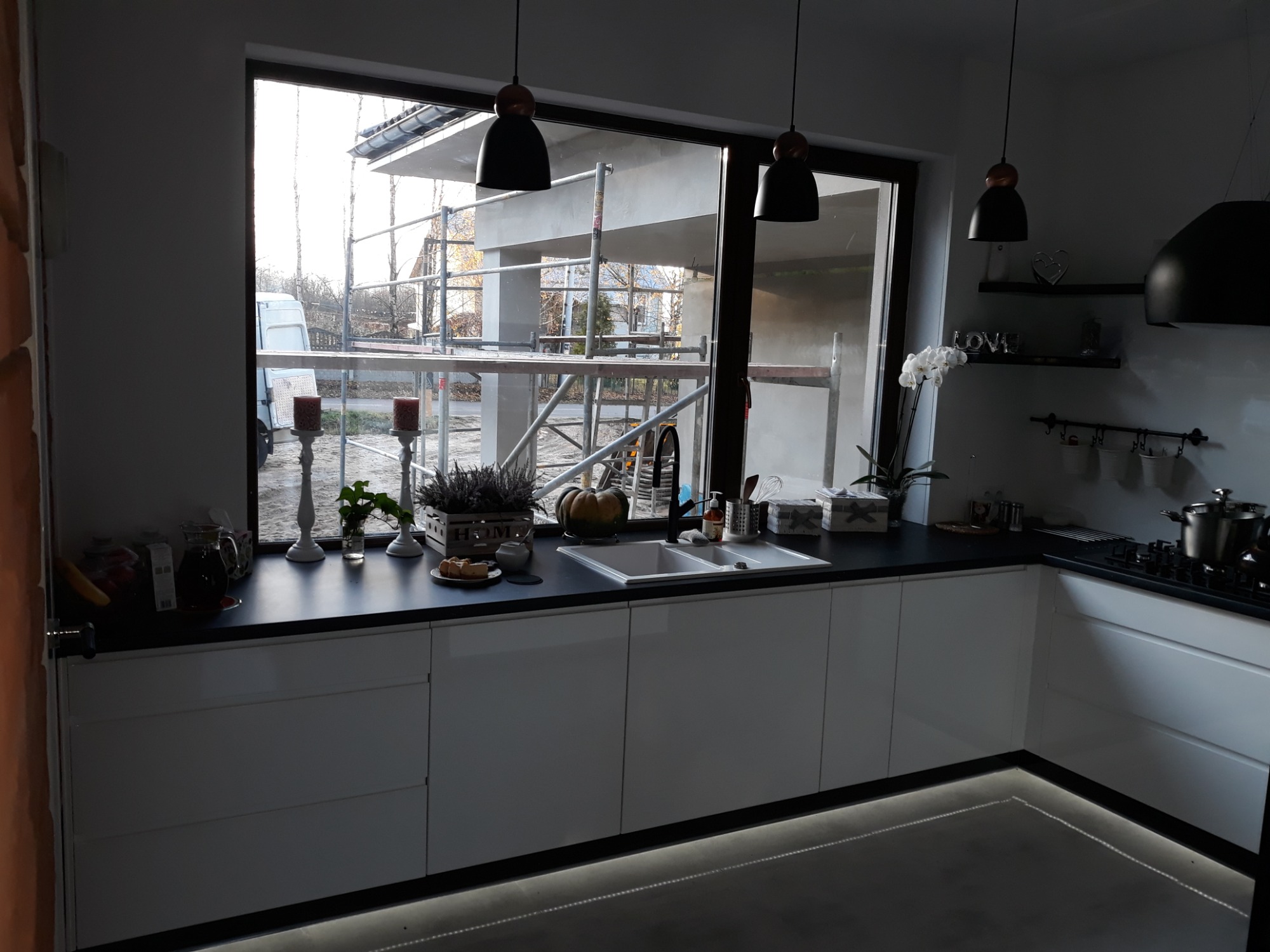Nowoczesna kuchnia z białymi szafkami i czarnym blatem, oświetleniem LED pod szafkami, widokiem na rusztowanie za oknem i dekoracjami na blacie.
