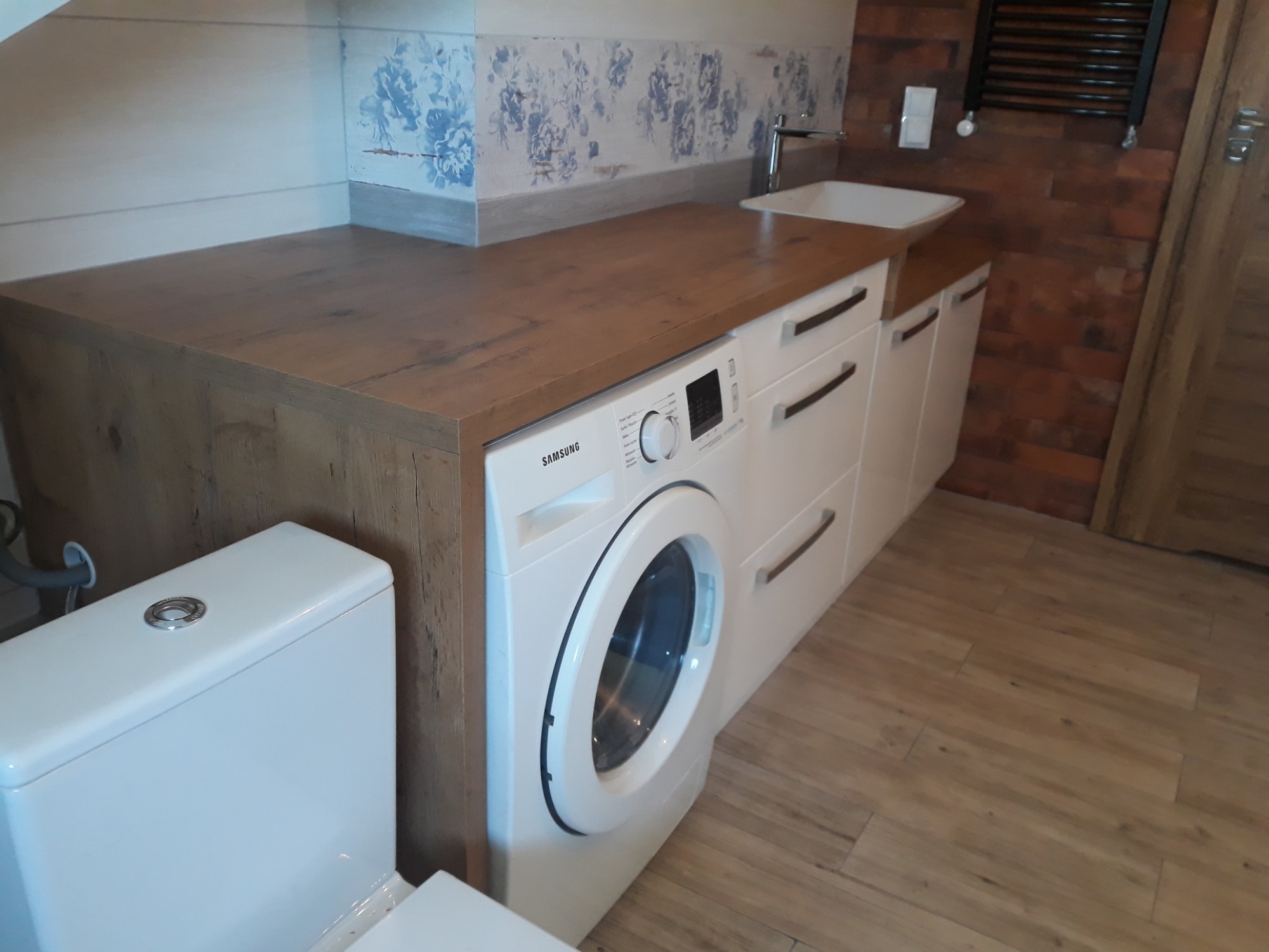 Nowoczesna łazienka z zabudową na wymiar: drewniany blat nad pralką i szafkami, umywalka nablatowa, toaleta kompaktowa, płytki ścienne z motywem kwiatowym i grzejnik drabinkowy na ścianie z cegły.