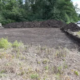 Przygotowany teren pod budowę, wykopane fundamenty z pryzmami ziemi, widok na ulicę z przejeżdżającymi samochodami i stosem drewna w tle.