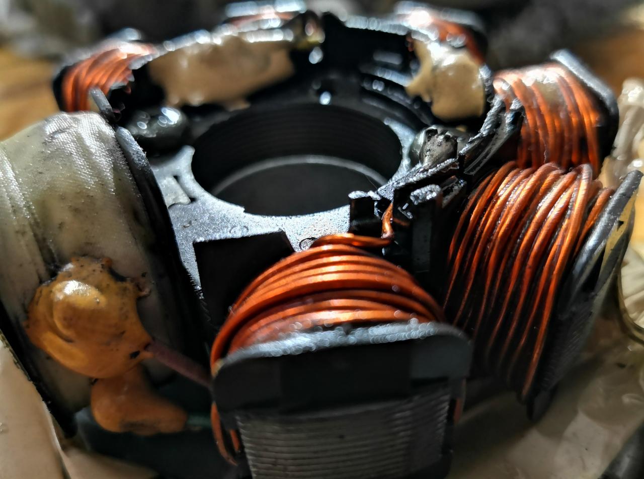 Zdemontowany, zabrudzony stojan alternatora samochodowego z widocznymi cewkami z miedzianego drutu i śladami oleju, sfotografowany z bliska.