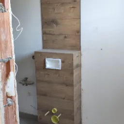 Obudowa stelaża WC wykończona panelami drewnopodobnymi, dopasowanymi do paneli ściennych i podłogi, z widocznymi przyłączami kanalizacyjnymi i fragmentem surowej ściany z cegły.