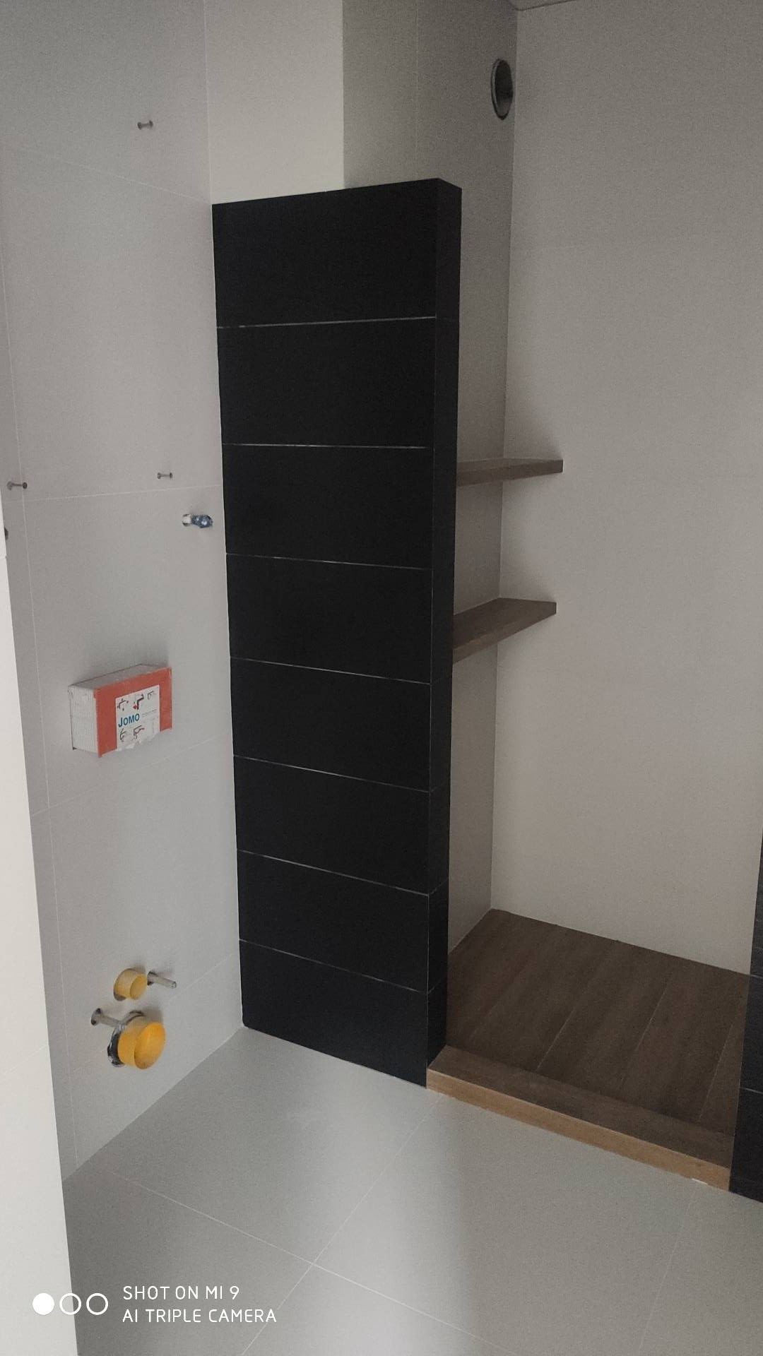 Fragment łazienki w trakcie remontu: widoczne białe płytki na ścianie, czarna obudowa wanny lub prysznica z poziomymi paskami, fragment podłogi wyłożonej płytkami imitującymi drewno, niezabudowane...