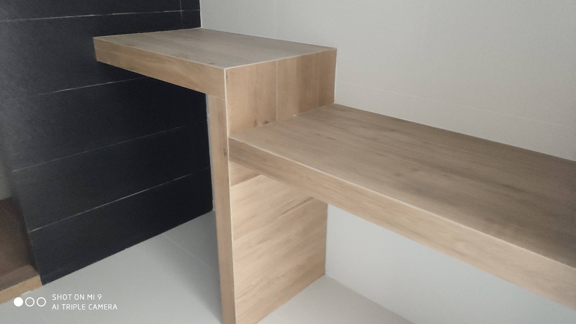 Półka z płytek ceramicznych imitujących drewno, zamontowana w łazience z czarnymi płytkami na ścianie, widok z dołu.