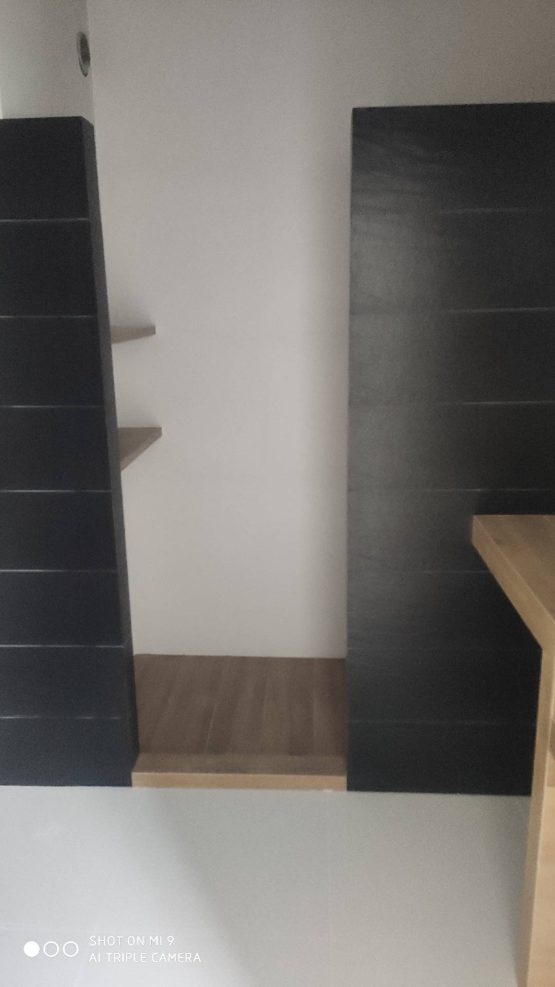 Fragment łazienki z czarnymi, poziomymi płytkami na ścianie i drewnianą podłogą, widoczne półki.