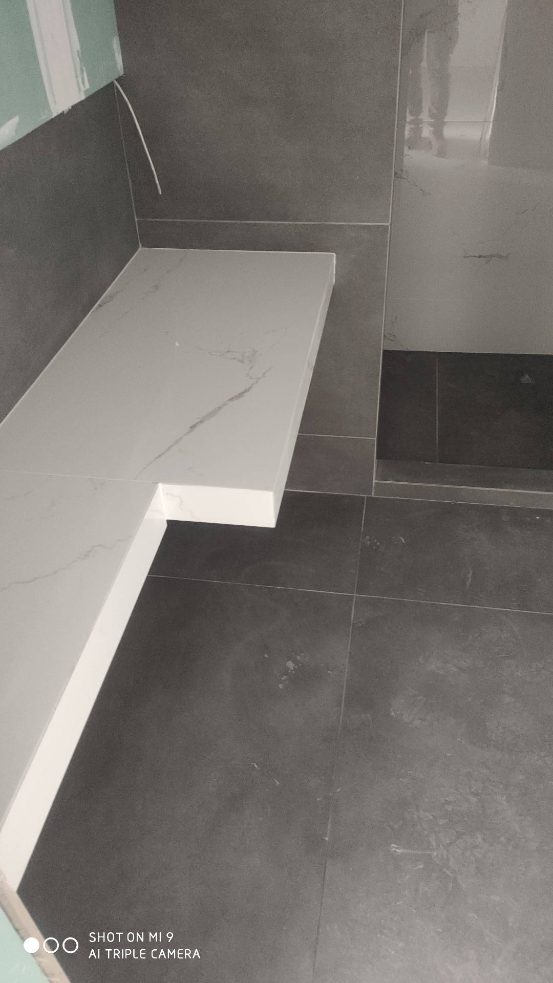 Wykończona łazienka z szarą glazurą i blatem z białego marmuru, widoczne płytki podłogowe i fragment ściany z niedokończonym wykończeniem.