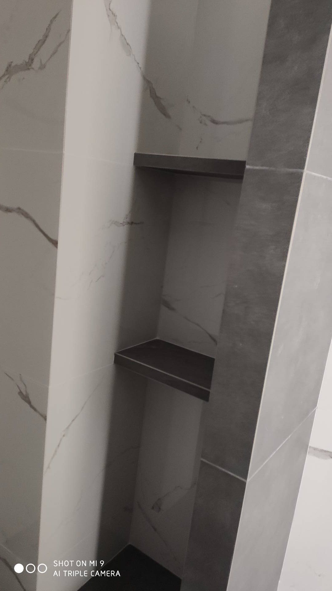 Wnęka prysznicowa z dwiema półkami wykończona białymi i szarymi płytkami imitującymi marmur, widok z dołu.